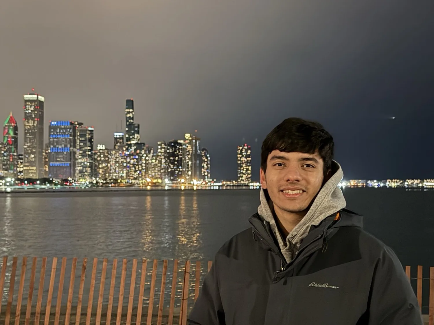 Uday Tyagi in a winter jacket in front of the Chicago skyline at night, city lights reflecting off the lake.