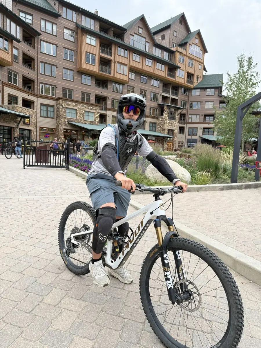 Uday in a full-face helmet and goggles, sitting on a mountain bike at the base of a mountain resort village.
