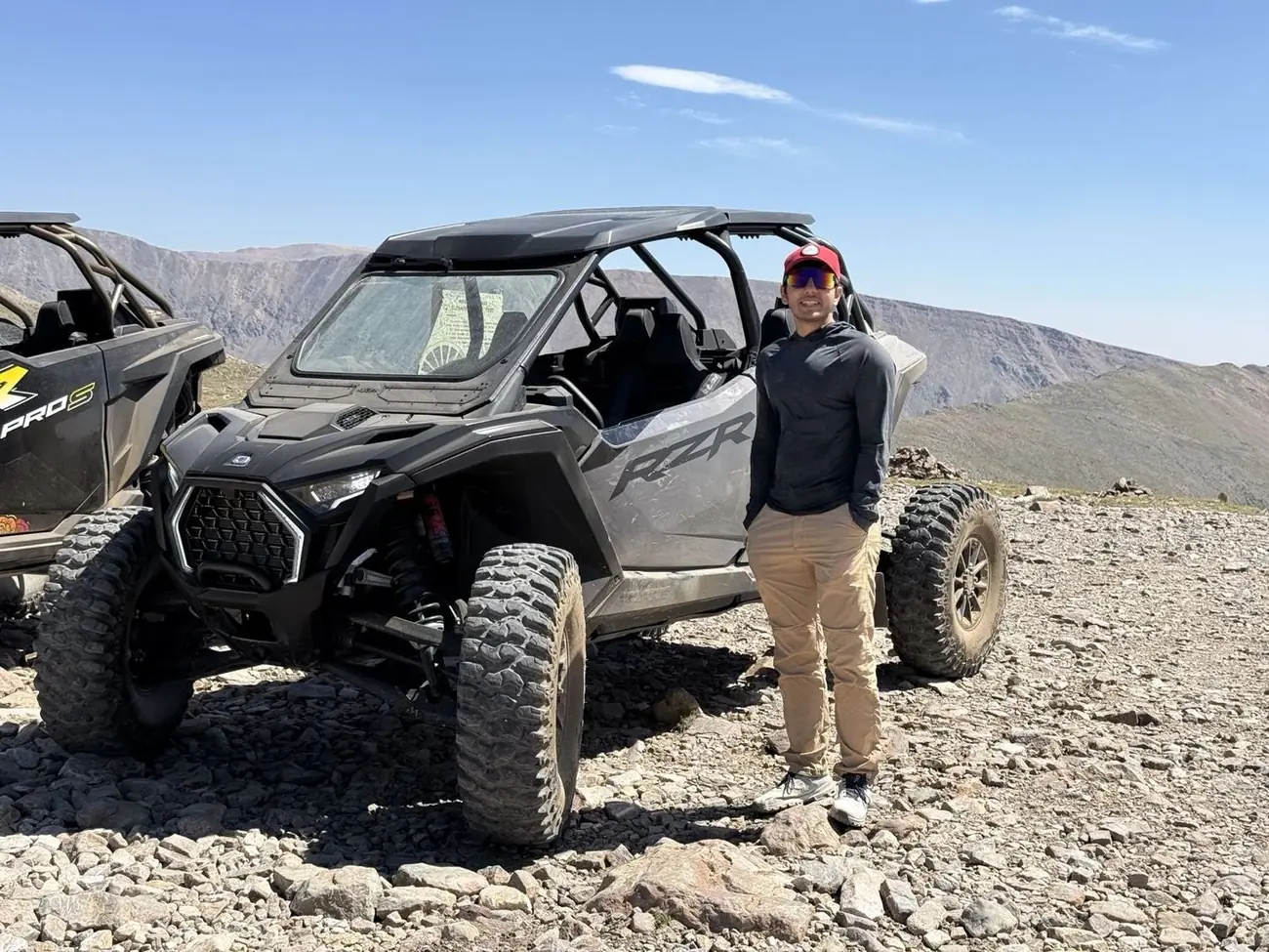 Uday standing beside a Polaris RZR off-road vehicle on a rocky ridge, with bare mountain slopes behind him.